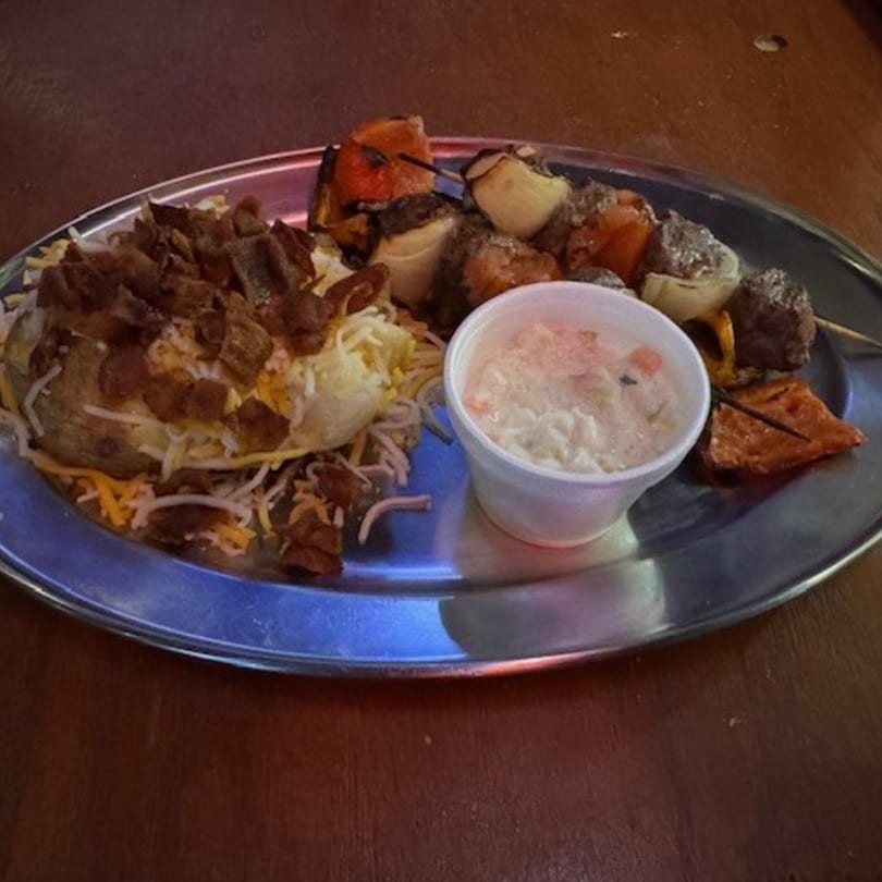 A loaded baked potato with bacon and cheese, a meat and vegetable skewer, and coleslaw on a metal plate.