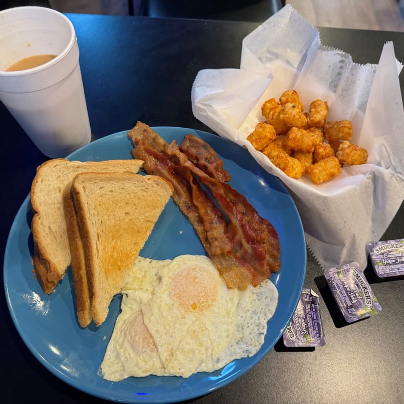A breakfast plate with bacon, fried eggs, toast, tater tots, and a cup of coffee.