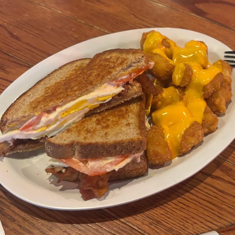 A breakfast sandwich with egg, bacon, and tomato on toast, served with cheese-covered tater tots.